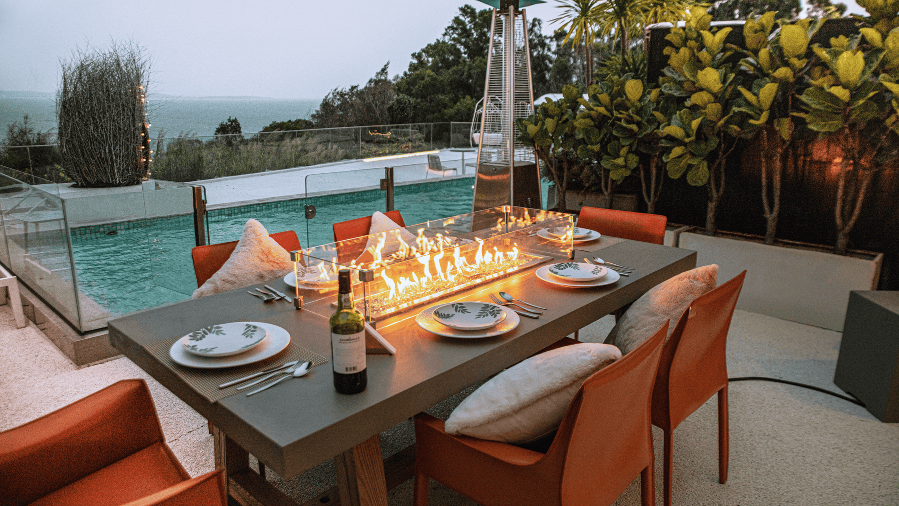 An outdoor dining table with built-in fire feature on a patio overlooking a pool.