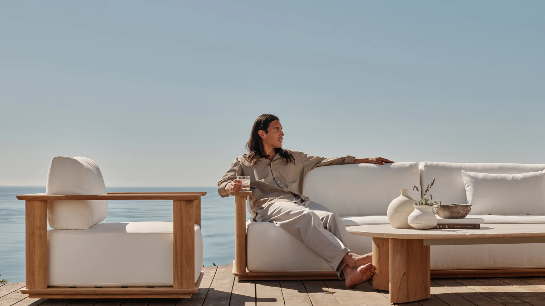 A man lounges outdoors on white upholstered outdoor sofa with a matching lounge chair near by and a view of the sea behind.