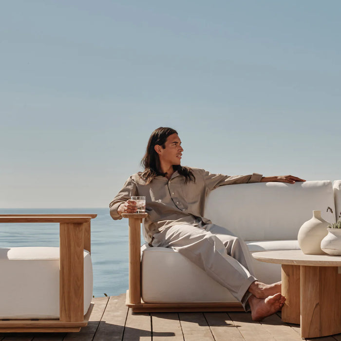 A man lounges outdoors on white upholstered outdoor sofa with a matching lounge chair near by and a view of the sea behind.