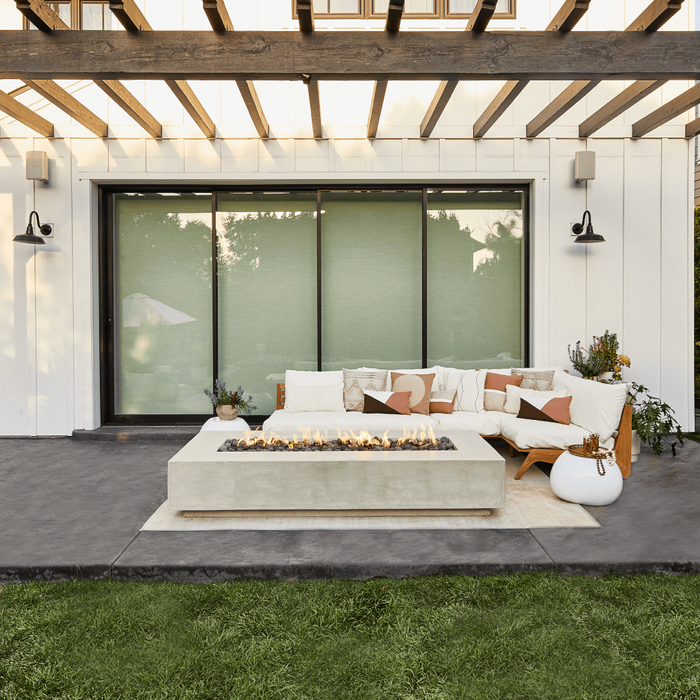 A modern outdoor patio with concrete fire pit, lounge sofa, and large potted plants all covered by a wooden pergola.