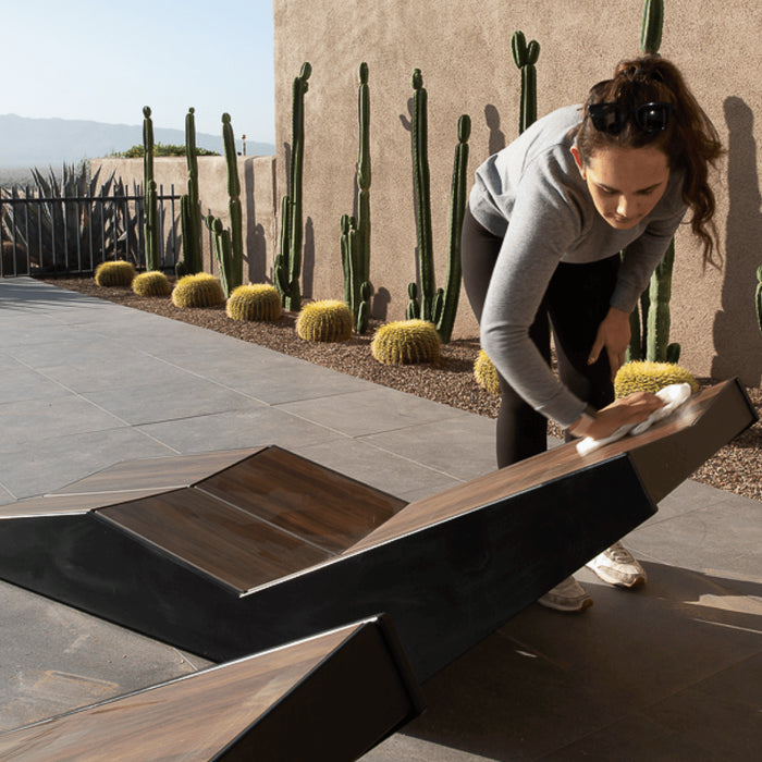 A woman cleans a range chaise lounger on a pool deck in front of a line of cactus plants