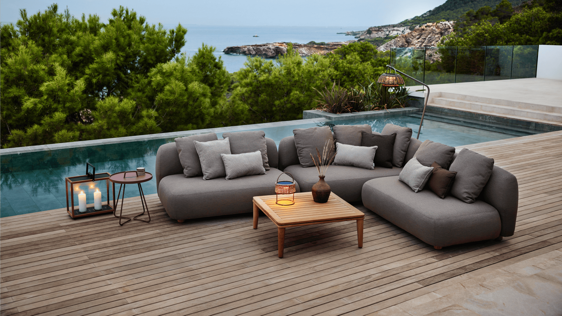 An outdoor lounge area beside a pool is illuminated with outdoor candles and a woven table lamp.