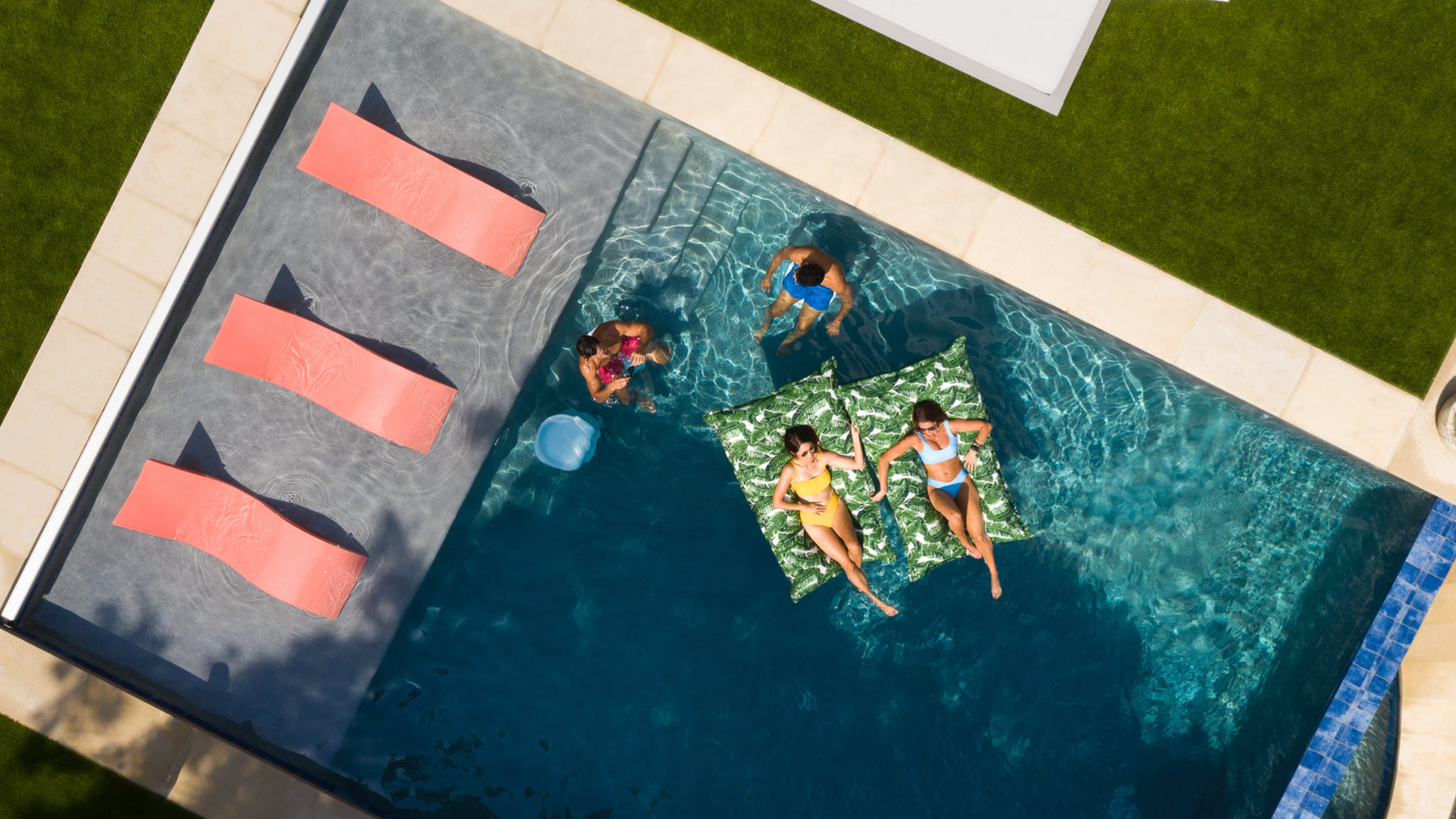 Three pink in-pool loungers on a tanning ledge while people float on in-pool pillows.