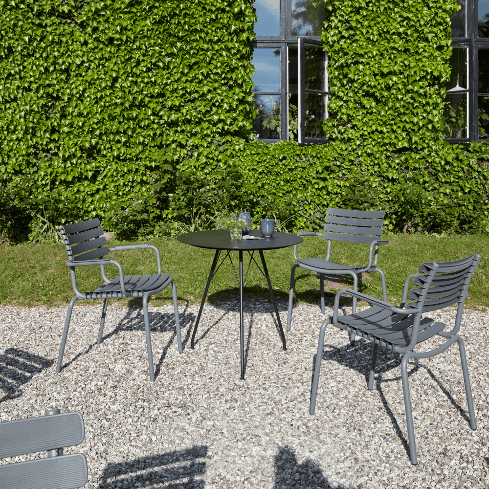 A black round outdoor coffee table and several gray outdoor dining armchairs set on a gravel surface beside greenery