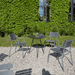 A black round outdoor coffee table and several gray outdoor dining armchairs set on a gravel surface beside greenery