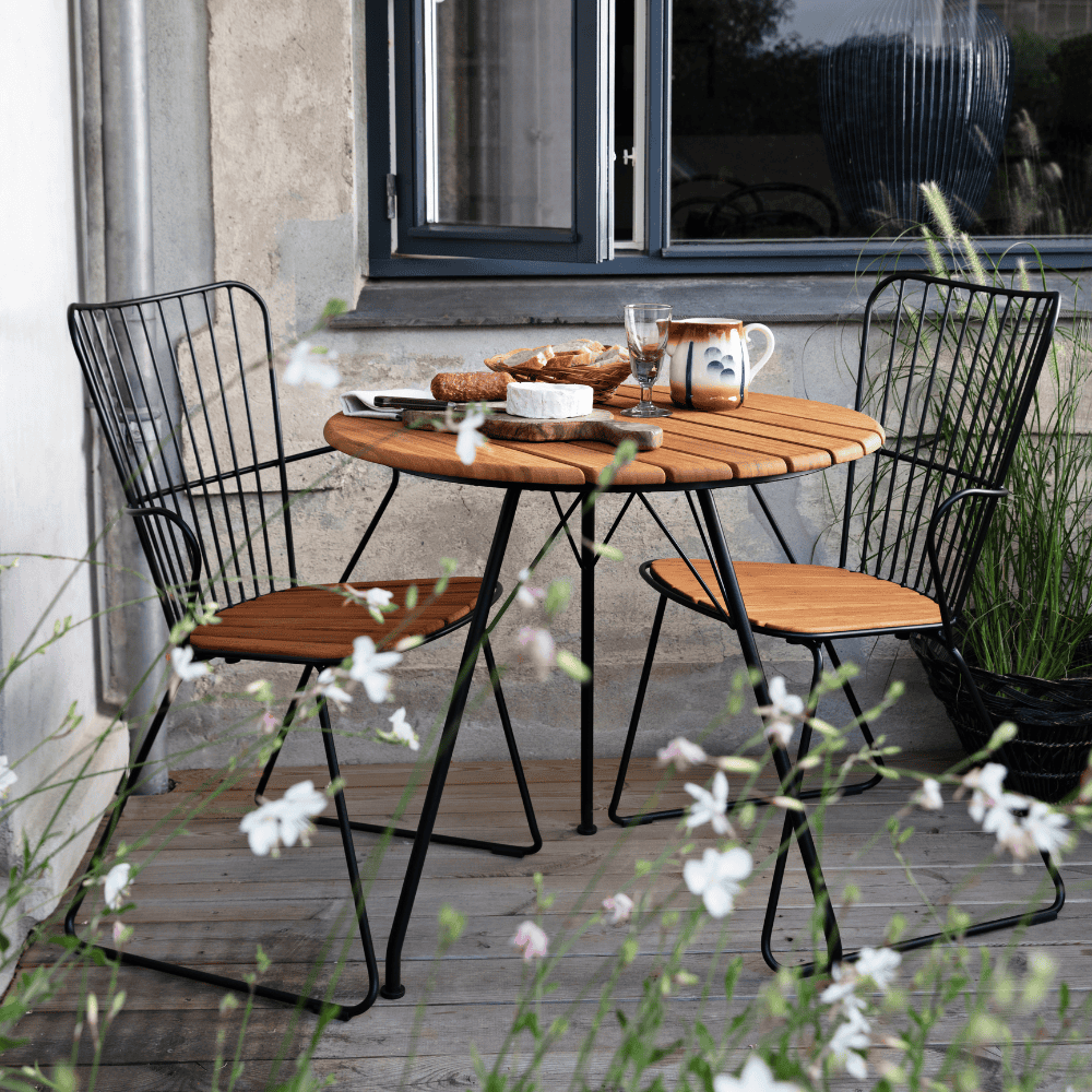 A round outdoor coffee table with bamboo lamella top and 2 outdoor dining armchairs with bamboo lamella seat set on a wooden deck patio