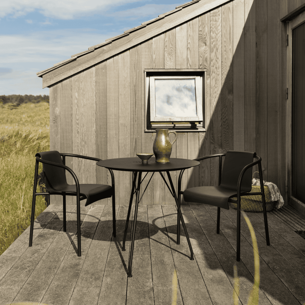 A black round outdoor coffee table and 2 black outdoor dining armchairs placed on a wooden deck patio beside small window