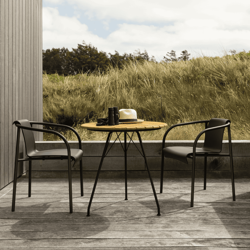 A round outdoor coffee table with bamboo lamella top and 2 black out dining armchairs set on a wooden deck near grassy field