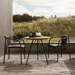 A round outdoor coffee table with bamboo lamella top and 2 black out dining armchairs set on a wooden deck near grassy field