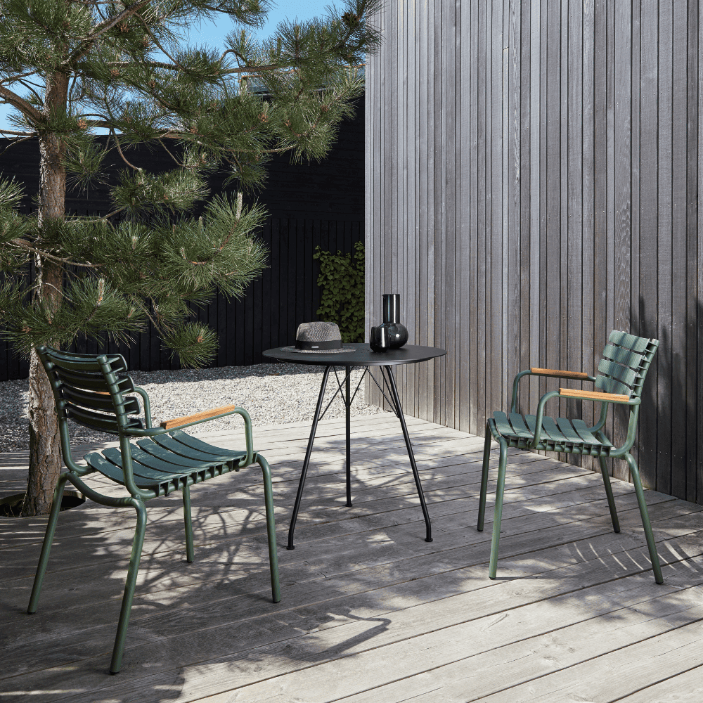 A black round outdoor coffee table and 2 green outdoor dining armchairs placed on a wooden deck patio beside the tree