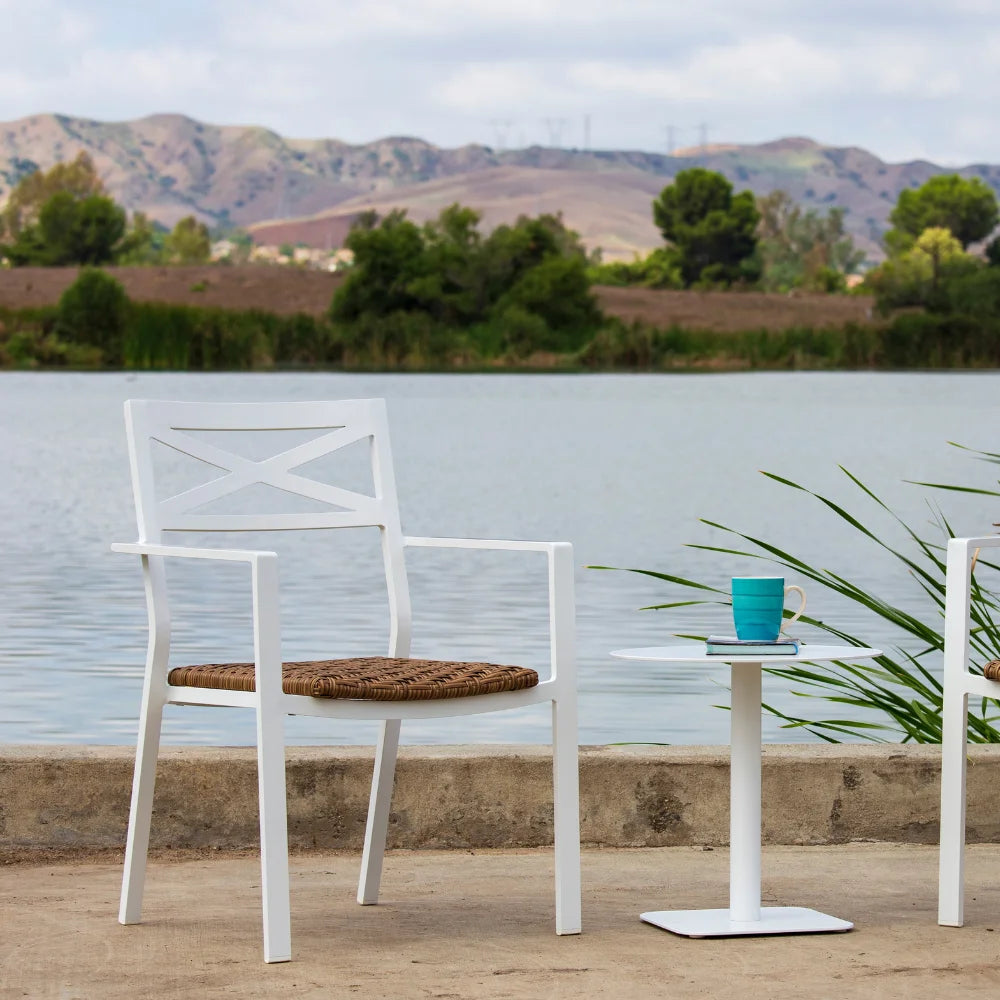 A white aluminum stackable outdoor dining armchair with padded wicker and a white round side table placed on a concrete surface near a body of water