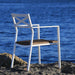 A white aluminum stackable outdoor dining armchair with padded wicker placed on a rocky surface by the water