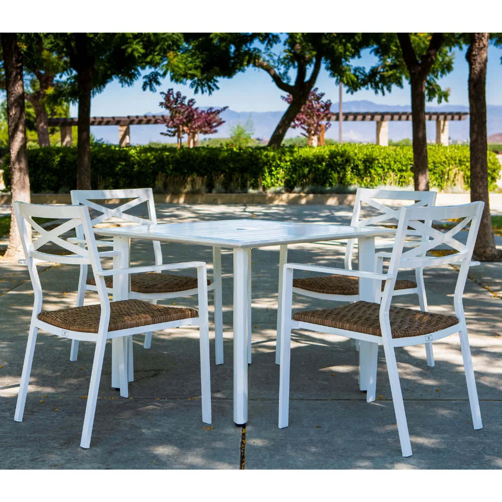 Four white aluminum stackable outdoor dining armchairs with white square dining table placed on an outdoor setting surrounded with plants and trees