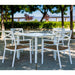 Four white aluminum stackable outdoor dining armchairs with white square dining table placed on an outdoor setting surrounded with plants and trees