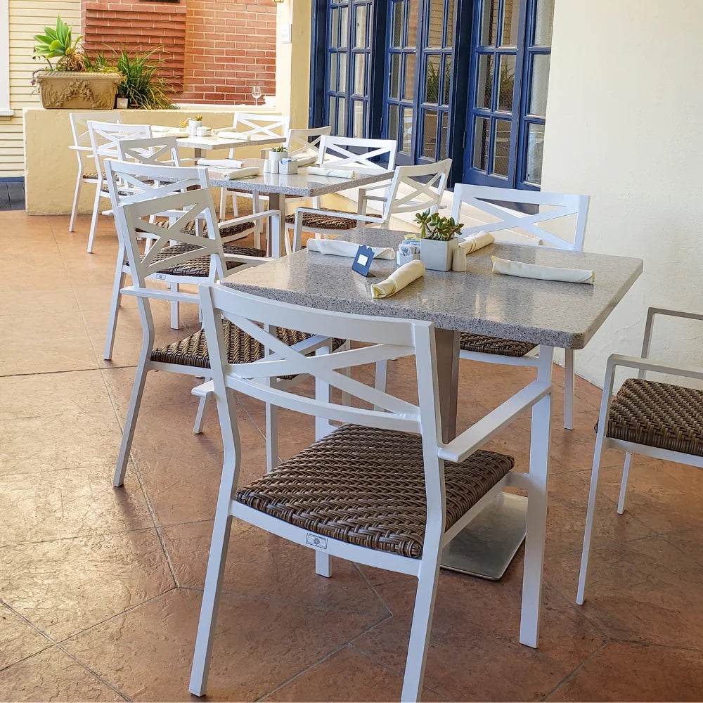 An outdoor dining area with multiple set of white aluminum stackable outdoor dining armchairs with padded wicker and square dining tables