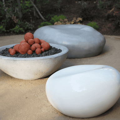 White fiberglass pebble garden seat and light grey concrete pebble garden seat on a sandy surface with grey concrete fire bowl 