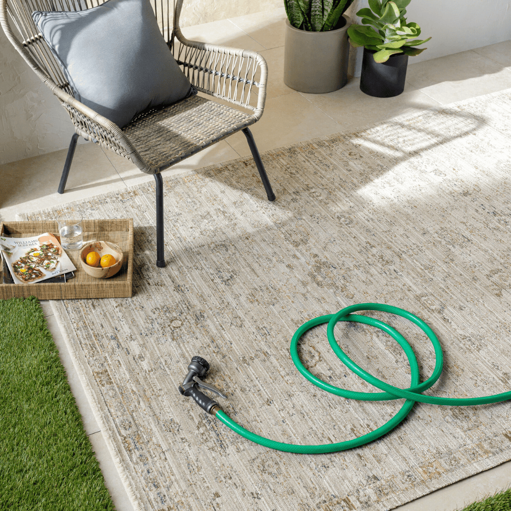 light brown rectangular machine woven outdoor rug with geometric pattern set on a patio with green hose and a woven chair