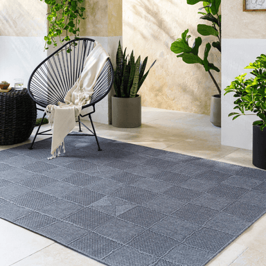 A dark gray checkered rectangular machine woven outdoor rug set on a patio with black acapulco chair and a variety of potted plants 