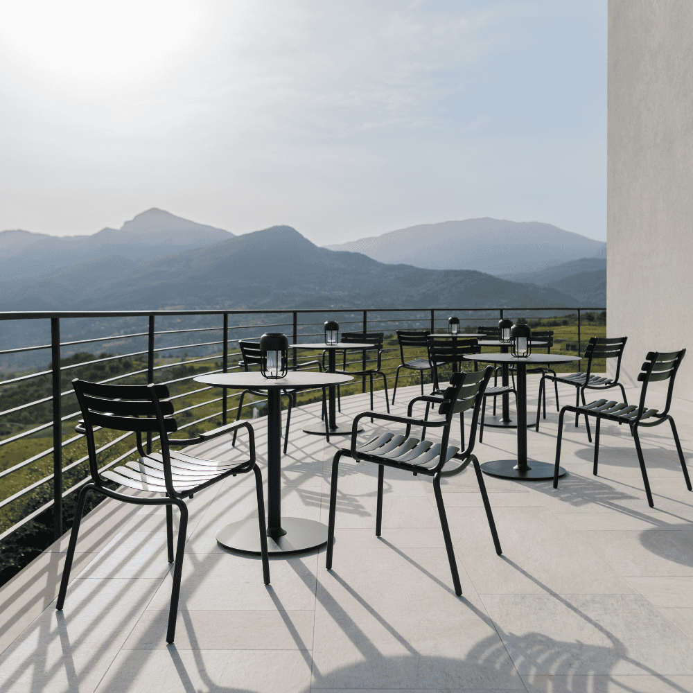 Several black stackable outdoor dining chairs and round outdoor dining tables set on a patio with black railings