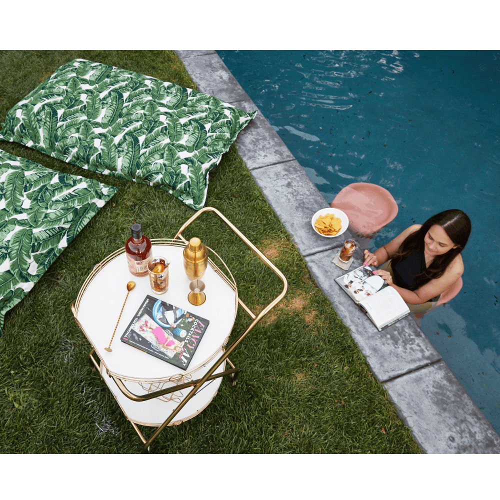 green white pool pillow on a grassy area beside the pool with a woman sitting on a pool barstool reading a magazine