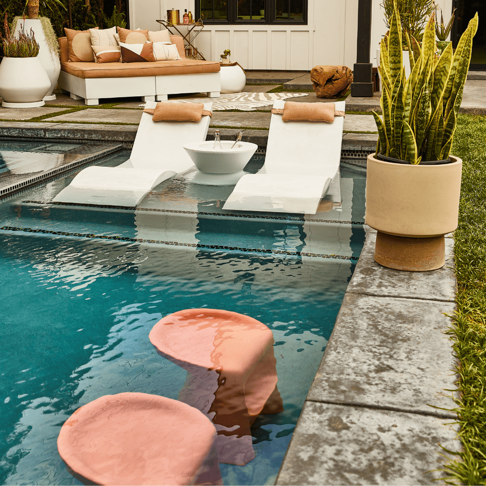 Pool barstool in a coral color placed in a pool together with 2 white pool chaise lounge and large sun bed