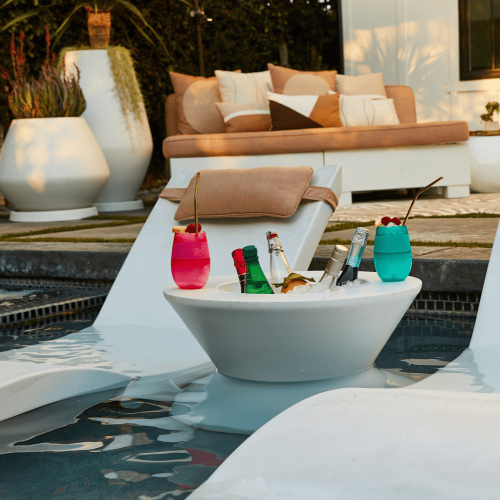 White tall ice bin side table with bottles of wine and cocktail on it placed in a pool in between 2 white pool chaise lounge