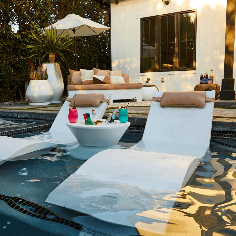 White tall ice bin side table with bottles of wine and cocktails on it placed in between 2 white pool chaise lounge