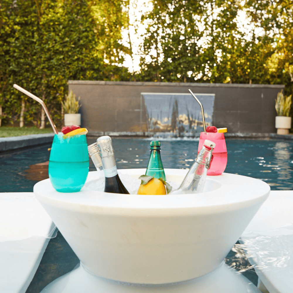 White tall ice bin side table with bottles of wine and cocktails on it placed in a pool