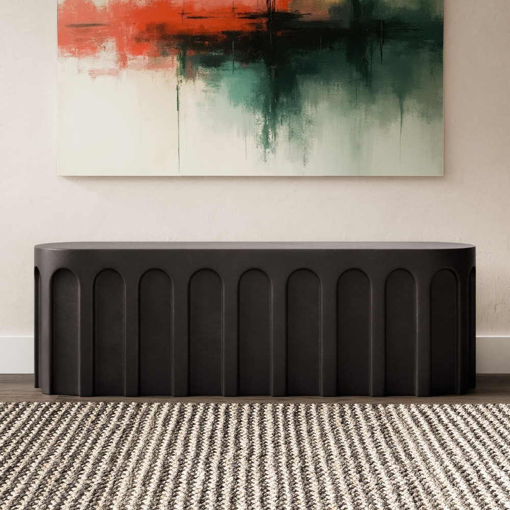 A matte black outdoor concrete oval bench with arched design pattern set in a living room against white wall with abstract painting