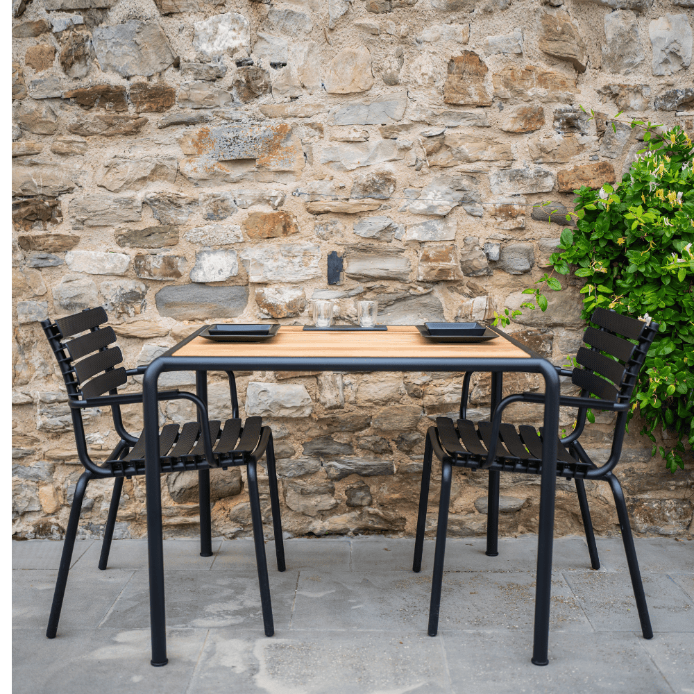 A square wooden outdoor dining table with black metal frame and 2 black outdoor dining chairs set on a patio against stone wall