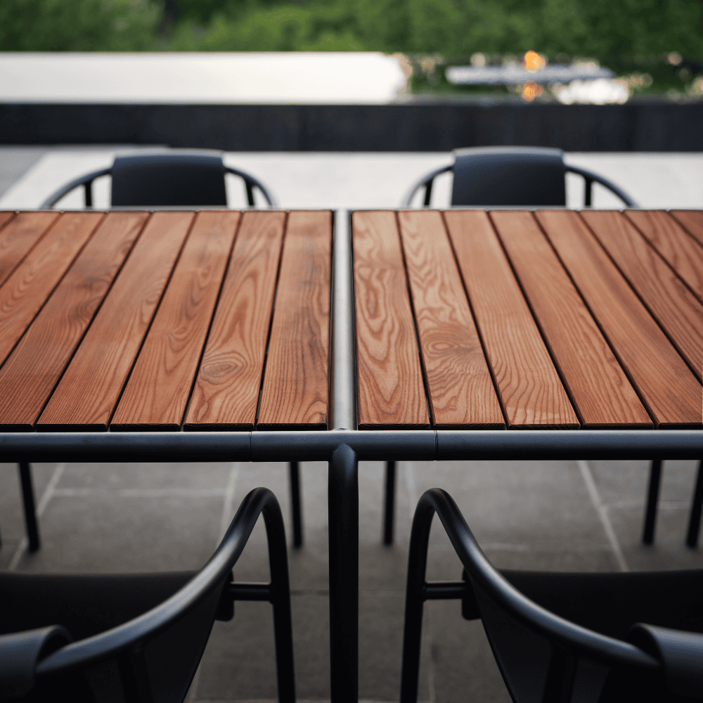 A wooden outdoor dining table with black metal frame set on a patio surrounded by 4 black outdoor dining chairs