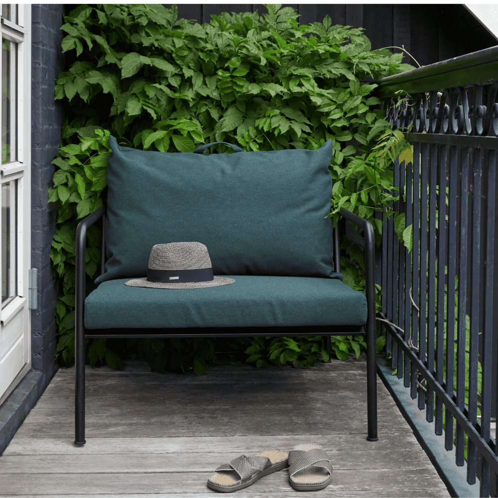 A dark green outdoor lounge chair with black aluminum frame set on a wooden deck balcony with black railings and plants