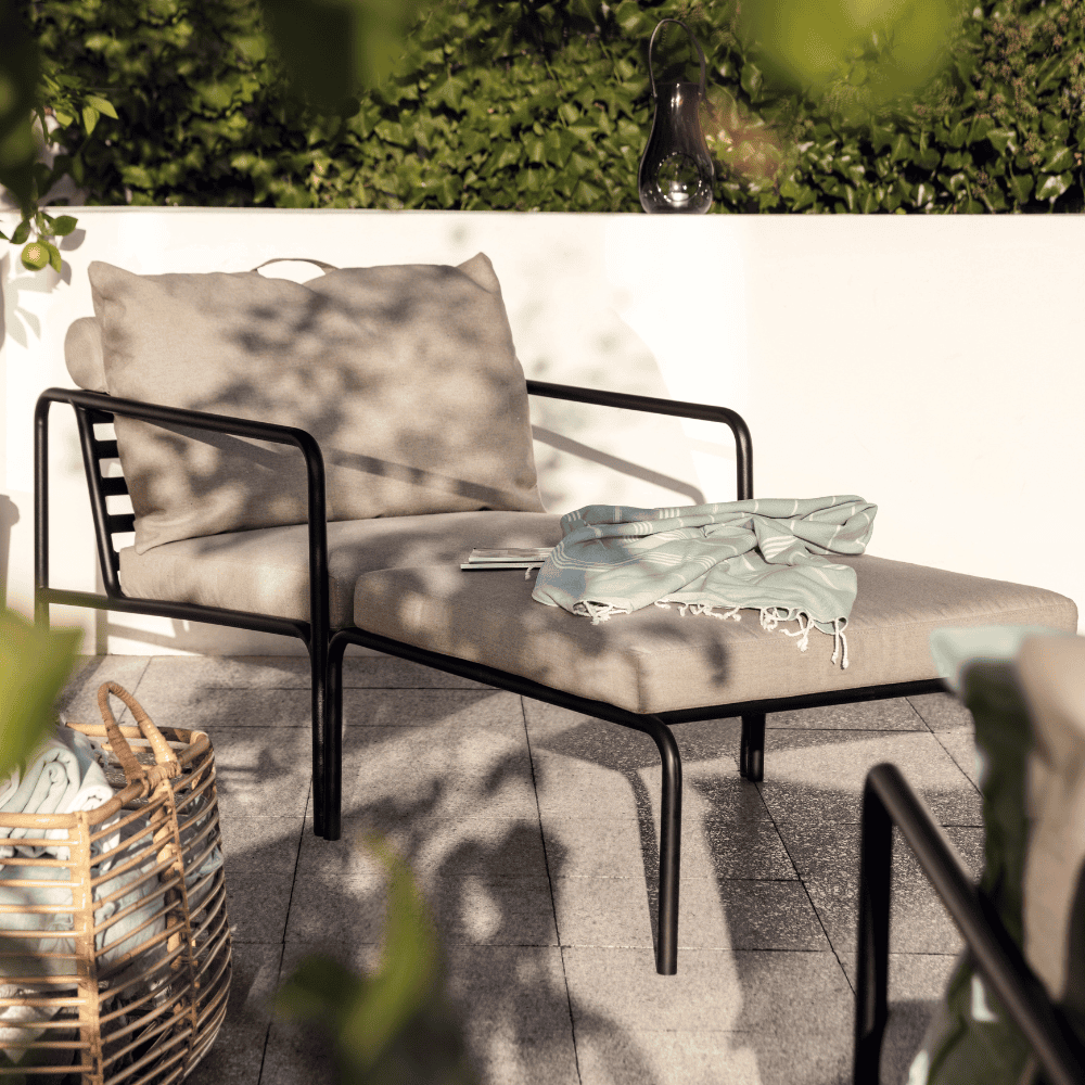 A beige outdoor lounge chair and outdoor ottoman with black aluminum frame set on a patio against white concrete fence