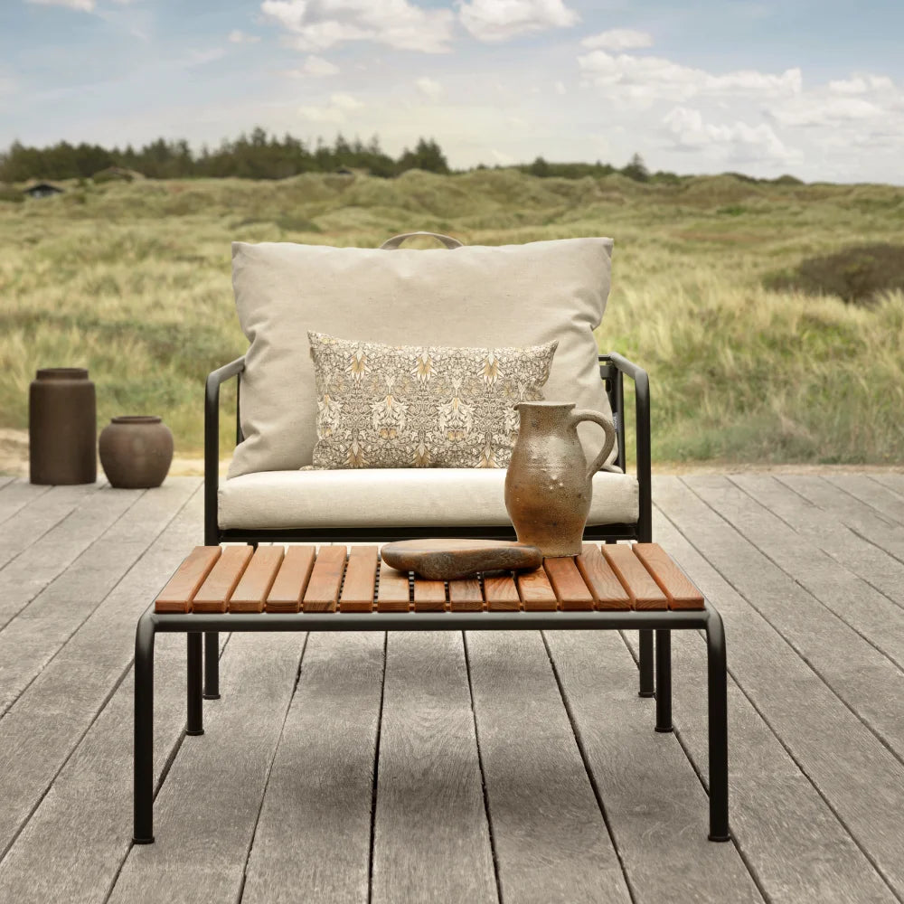 A beige outdoor lounge chair with black aluminum frame and a wooden outdoor lounge table set on a patio near grassy field