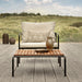 A wooden outdoor lounge table with black metal frame and a beige outdoor lounge chair set on a wooden deck near grassy field