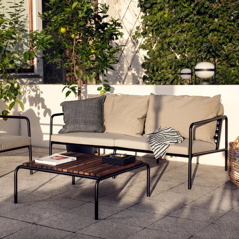 A wooden outdoor lounge table with black metal frame and a beige outdoor lounge chair set on a patio surrounded by plants