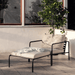 A beige outdoor ottoman and outdoor lounge chair with black aluminum frame set on a tiled patio beside glass window