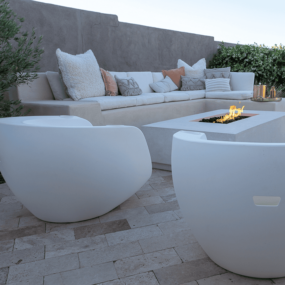 Two white moon Shaped pool chairs placed on a patio together with a concrete fire table and L shaped white outdoor Sofa