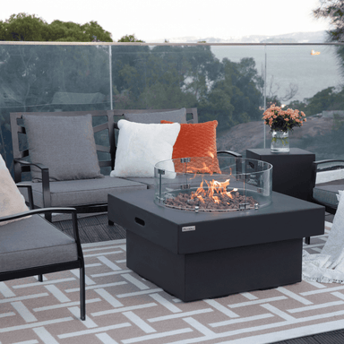 Black square concrete fire pit table with glass win screen placed on a patio surrounded by grey sofa and chairs