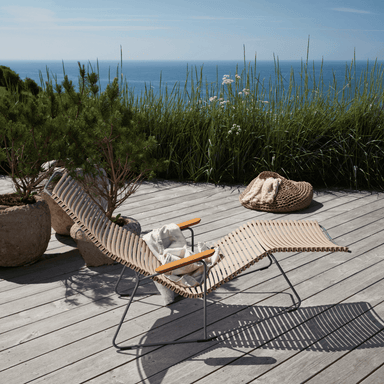 A light brown outdoor sunrocker with bamboo armrest placed on a wooden deck surrounded by potted plants and tall grass