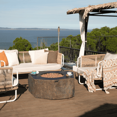 Brown hexagonal shaped outdoor concrete fire pit table surrounded by sofa and chairs set on a wooden deck patio with serene view of the sea