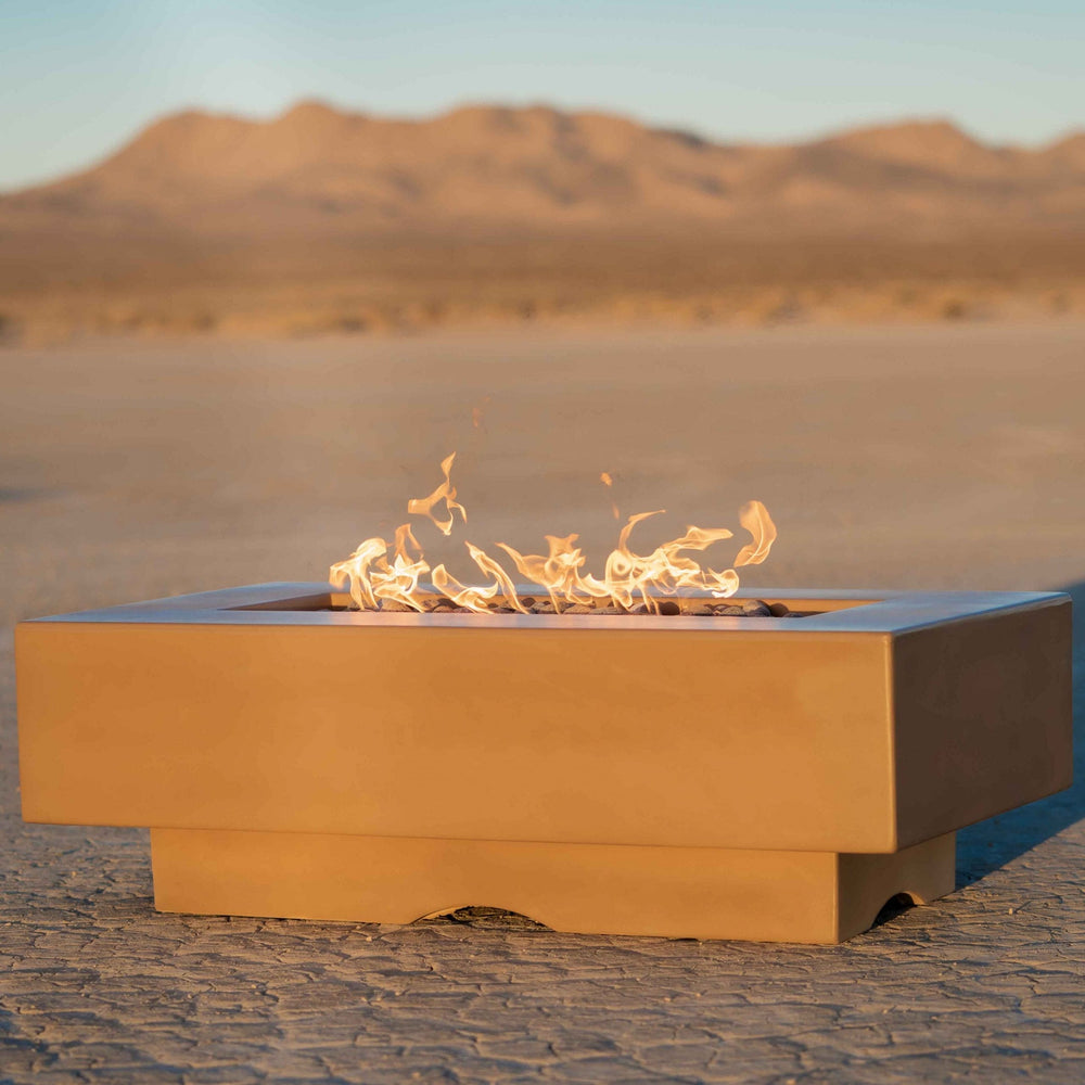 A brown outdoor square concrete fire pit table with flames set on a cracked dry desert ground with a view of a distant hill