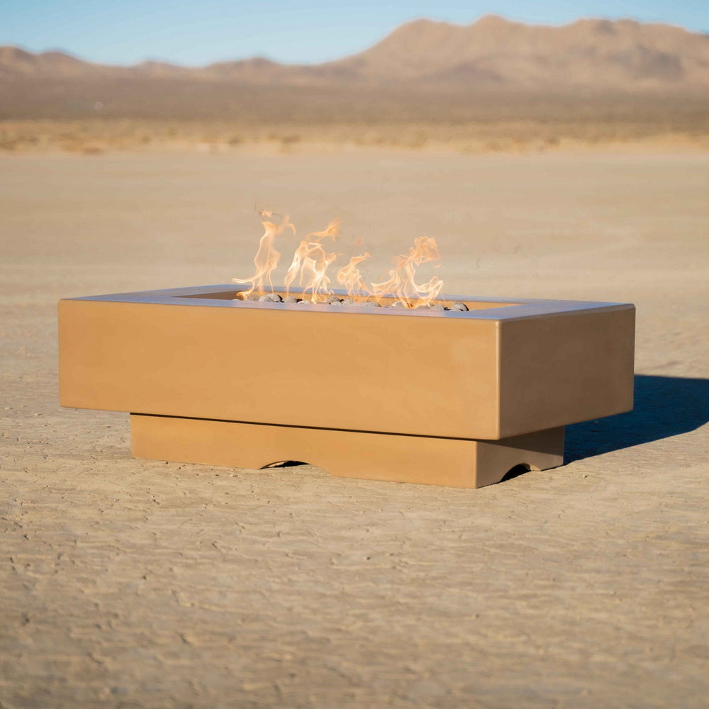 A brown outdoor square concrete fire pit table with flames set on a cracked dry desert ground with a view of a distant hill