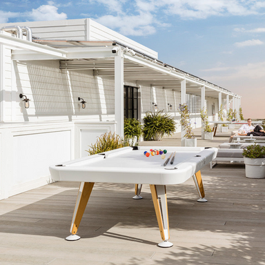 A white rectangular outdoor pool table set on a wooden deck outdoor patio with several potted plants