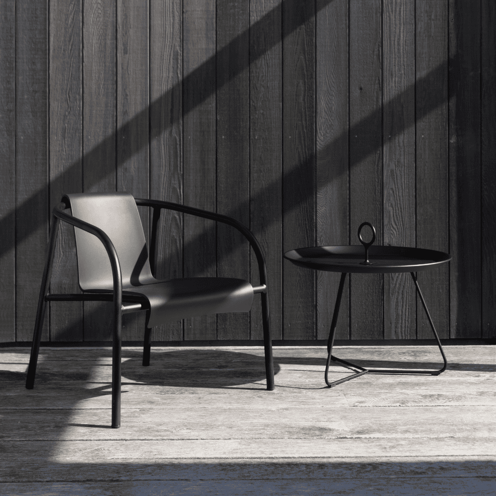 A black round outdoor tray table and a black metal outdoor lounge chair set on a wooden deck against wooden wall