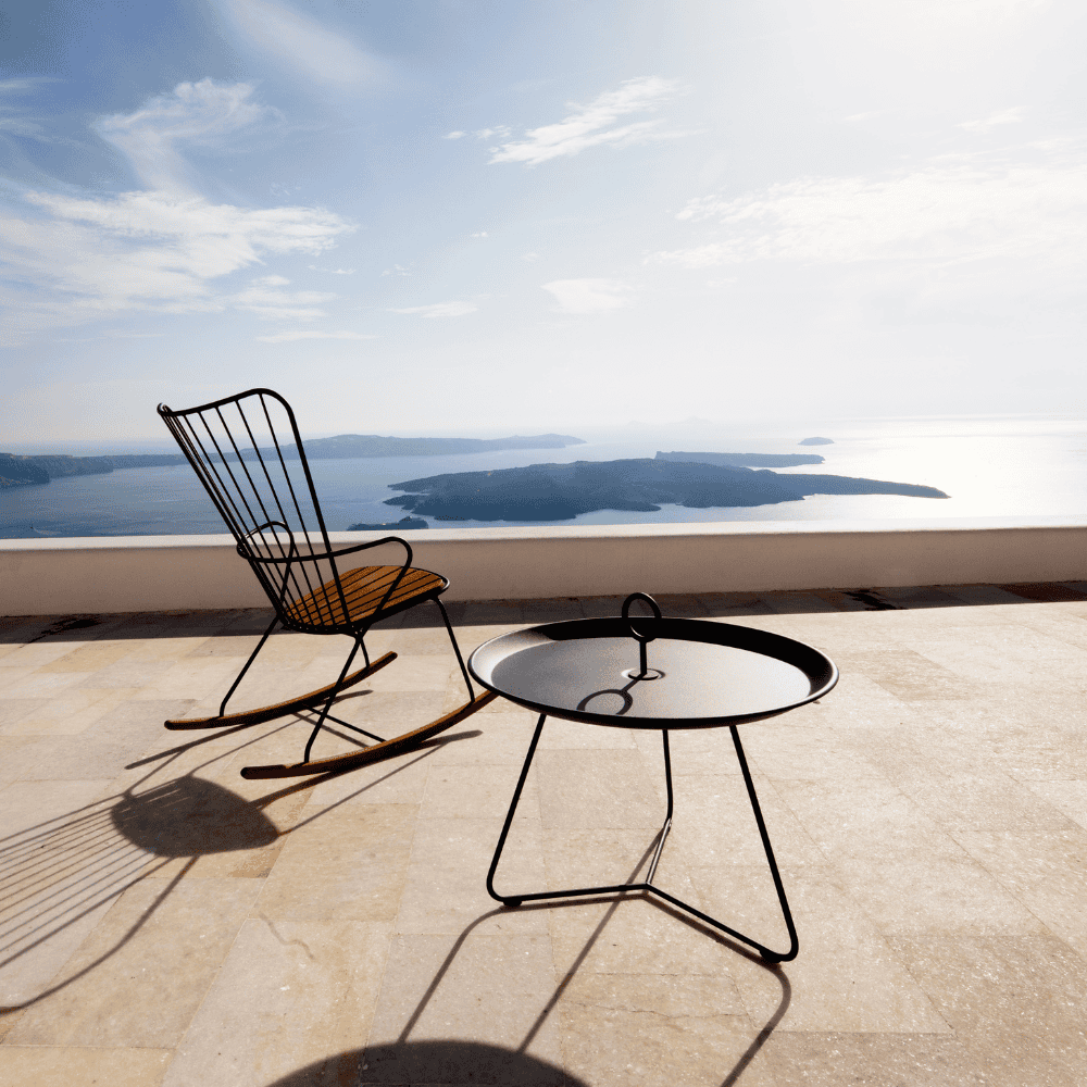 A black round outdoor tray table and an outdoor rocking chair set on a tiled patio with scenic view of a sea and islands