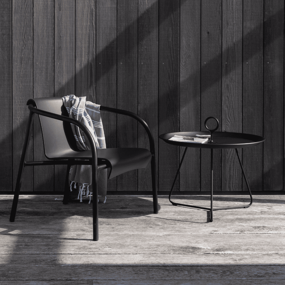 A black round outdoor tray table and a black metal outdoor lounge chair with gray fabric on it set on a wooden deck patio