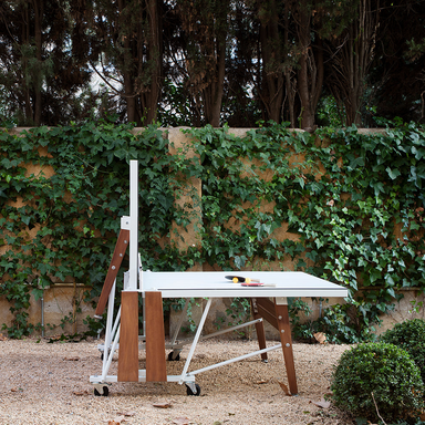 A white folding outdoor ping pong table set on a gravel surface beside a wall with lush green ivy