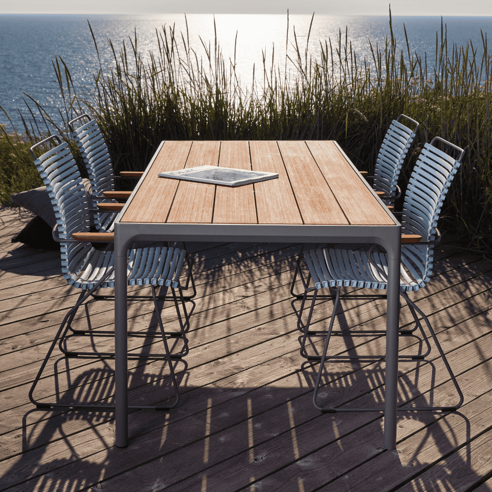 A rectangular wooden outdoor dining table with gray aluminum frame and 4 white dining chairs set on a wooden deck with tall grass