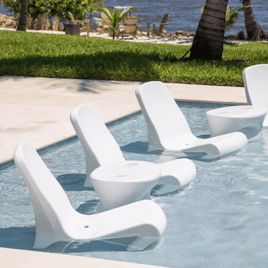 four white pool chairs and two white pool side table in a pool beside grassy field.